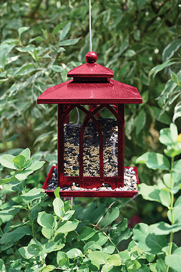 Burgundy rectangle shaped feeder with a pyramid shaped lid There are windows on each side to see the seeds It is hanging outdoors.