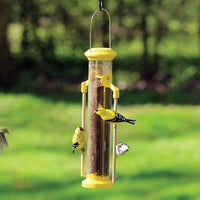 Feeder is hanging outdoors while 3 goldfinches are feeding while holding onto the perch, and one more goldfinch is flying towards the feeder.