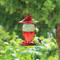 Feeder is hanging outdoors on a tree. It is filled halfway with nectar. A hummingbird is flying to drink from the feeding hole.