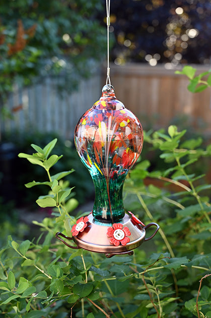 Circle shaped glass with red, yellow, orange, and green markings on it. Copper coloured base with red flower shaped feeding holes.