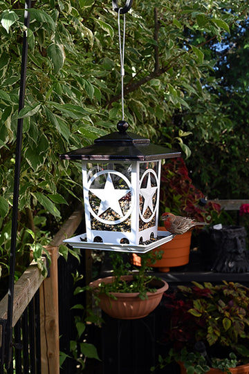 White feeder with star designs on each side. Black roof. It is filled halfway with seeds, and hanging from a pole system outdoors.