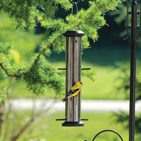 Feeder is hanging from a pole outdoors, the feeder is filled with seed while a gold finch sits on the perch for feeding. 