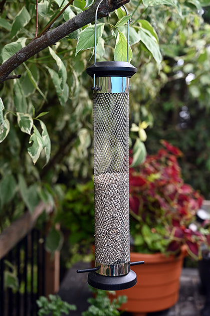 Hanging on a tree. The feeder is half-filled with seeds. Black lid, perches, and base. Metal tube mesh.