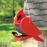 Feeder is hanging outdoors filled with seed while a grosbeak feeds.