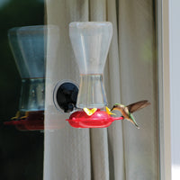 The hummingbird feeder is outside attached to a window while a hummingbird is enjoying the nectar.