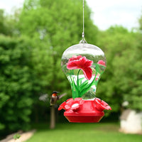 Feeder is hanging outdoors while a hummingbird is flying towards the nectar.