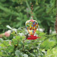 The feeder is hanging from a tree outdoors while a hummingbird is feeding.