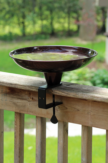 Bowl shaped bird bath that is brown in colour, outdoors with water inside the bird bath, on the deck railing.