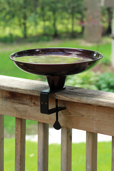 Bowl shaped bird bath that is brown in colour, outdoors with water inside the bird bath, on the deck railing.